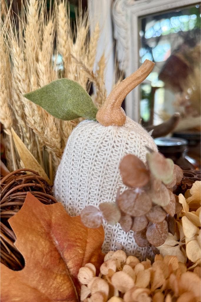 A white, knitted pumpkin with a fabric stem and leaf is surrounded by dried wheat, brown leaves, and beige hydrangea flowers, creating a cozy autumn-themed arrangement.