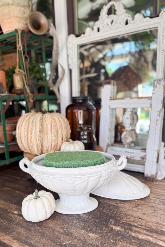 A white decorative bowl with handles holds a green floral foam block, placed on a rustic wooden table alongside small white pumpkins. Behind it are amber bottles, a vintage mirror, and textured décor.