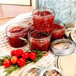 Five small jars of homemade tomato jam with lids beside them, sitting on a textured towel. In front, there’s a sprig of rosemary and a bunch of fresh cherry tomatoes—an inviting setup perfect for anyone following an Easy Cherry Tomato Jam Recipe for Beginners. A large metal container is in the background.