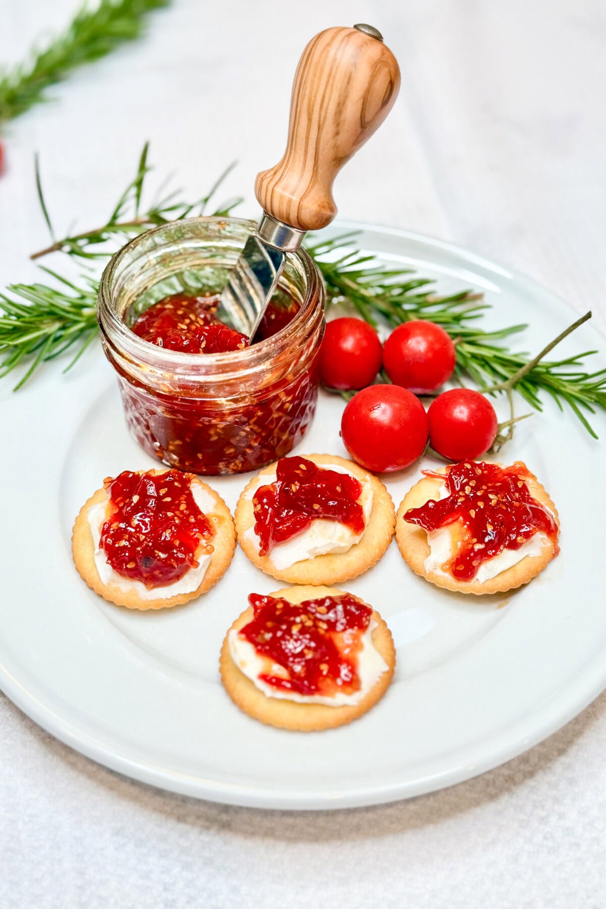 A plate with four crackers topped with cream cheese and red jam sits next to a jar of Easy Cherry Tomato Jam Recipe for Beginners, a wooden-handled spreader, fresh tomatoes, and sprigs of rosemary on a white surface.