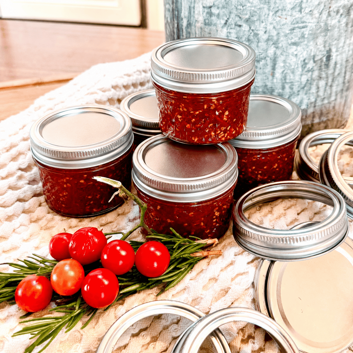 Five small jars filled with red homemade sauce are stacked on a towel, with rosemary and cherry tomatoes in front—perfect props to highlight an Easy Cherry Tomato Jam Recipe for Beginners. Extra jar lids are scattered nearby.