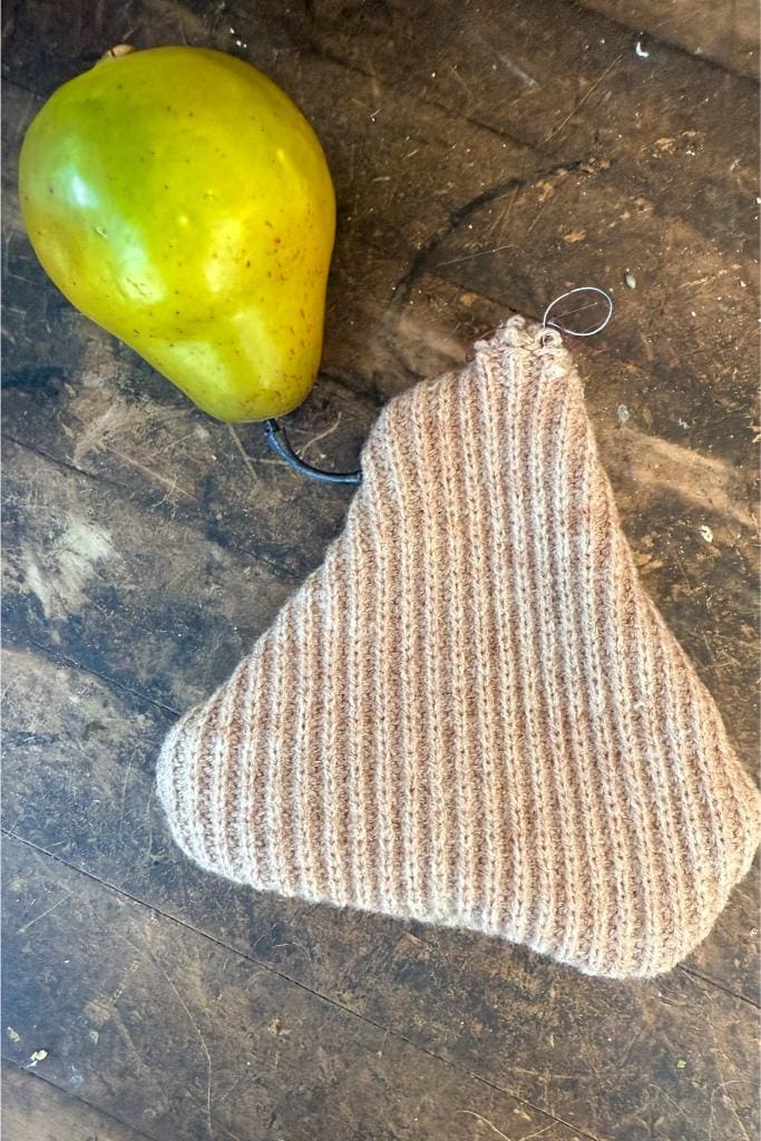 DIY Sweater Pears-A green pear is attached by its stem to a knitted, ribbed beige pouch, both resting on a worn wooden surface.