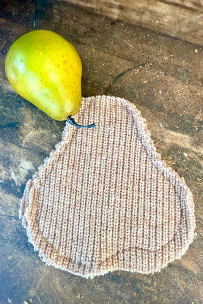 DIY Sweater Pears-A yellow-green pear placed next to a knitted pear-shaped coaster on a rustic wooden surface. The coaster is beige with a black string at the top to mimic a stem.