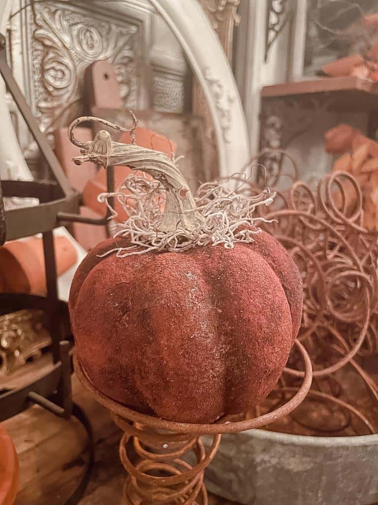 A plush, velvet pumpkin with a curly stem and decorative moss sits on a metal stand amid rustic, autumn-themed decor—perfect inspiration for your next pumpkin crafts project.
