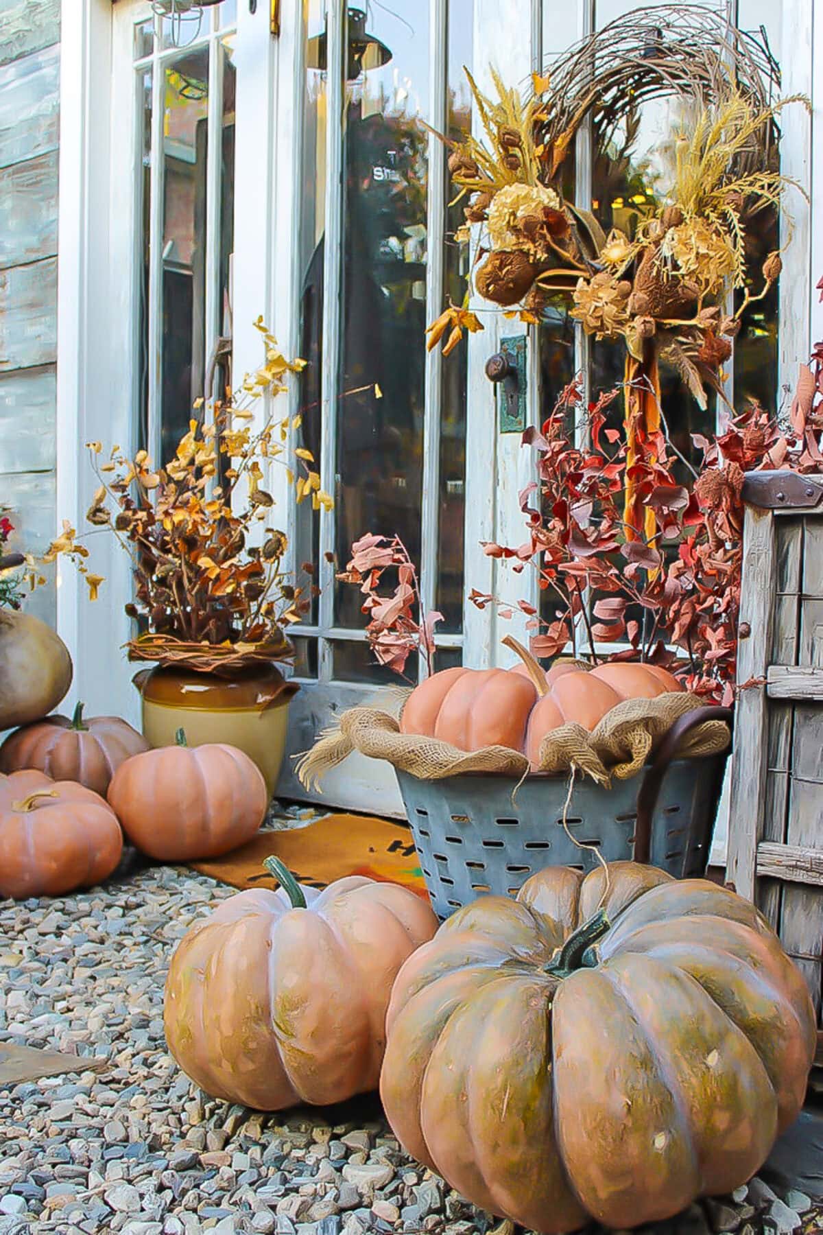 A fall porch display with pumpkins on gravel, potted plants with dried autumn leaves, and a wreath of dried flowers offers cozy she shed decorating ideas for fall, complete with a basket of pumpkins adorned with burlap bows.