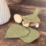 Green felt leaves and small wooden sticks arranged on a rustic wooden surface, with part of a knitted off-white object—resembling DIY Sweater Pears—visible on the left side of the image.