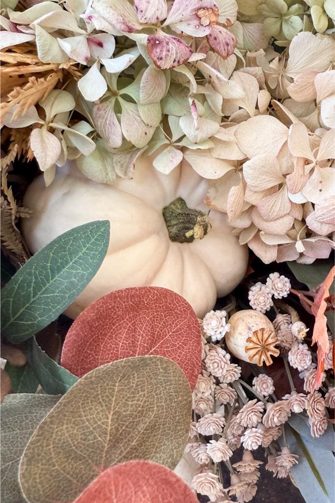 A white pumpkin surrounded by dried hydrangeas, poppy pods, and various green, red, and brown leaves arranged in an autumnal display.