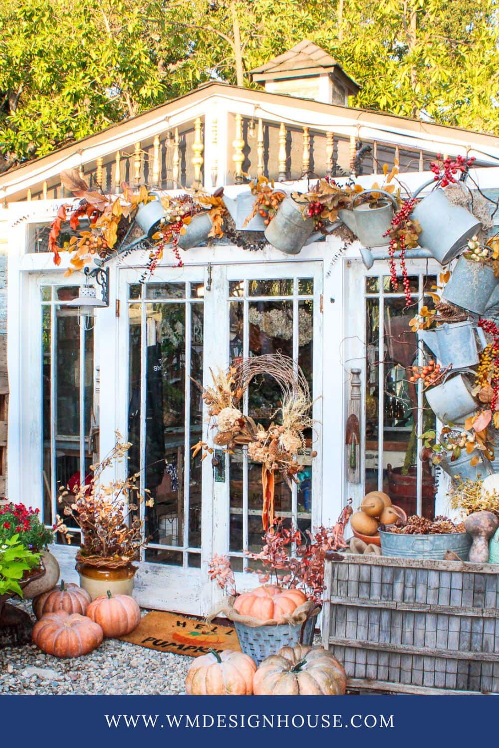 A garden shed with glass doors decorated for fall with metal pitchers, autumn leaves, pumpkins, and dried flowers. A wooden crate, potted plants, and various pumpkins are arranged on the gravel path in front.