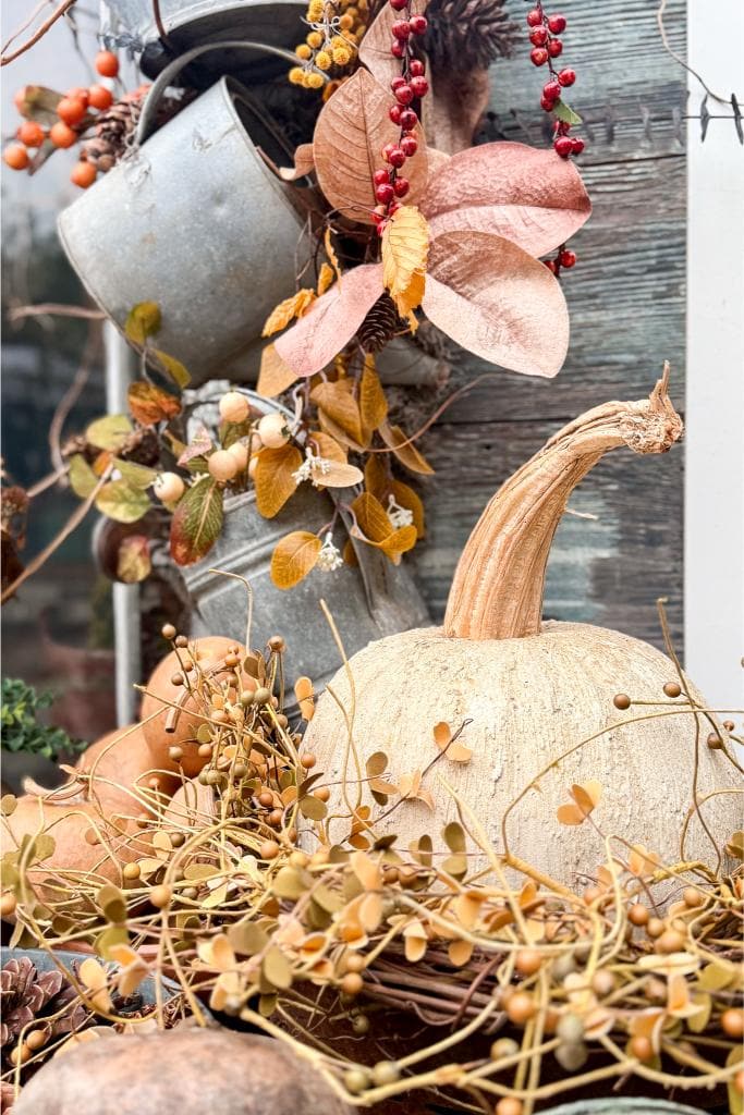 A pale pumpkin sits among dried leaves, berries, and twigs, with a rustic metal container behind it, creating an autumn-themed arrangement against a weathered wooden background.
