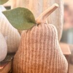 A decorative pear made of light brown knitted fabric with a felt leaf and a wooden stem, placed indoors with other soft-toned decor in the background.