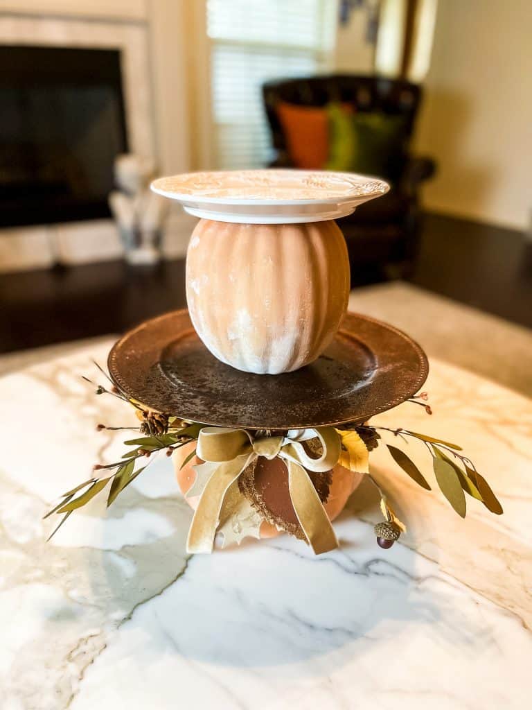 A cozy DIY pumpkin-shaped centerpiece—perfect for Pumpkin Crafts—made from stacked plates and bowls painted orange and white, adorned with ribbons, leaves, and acorns, sits on a marble table in a welcoming living room.