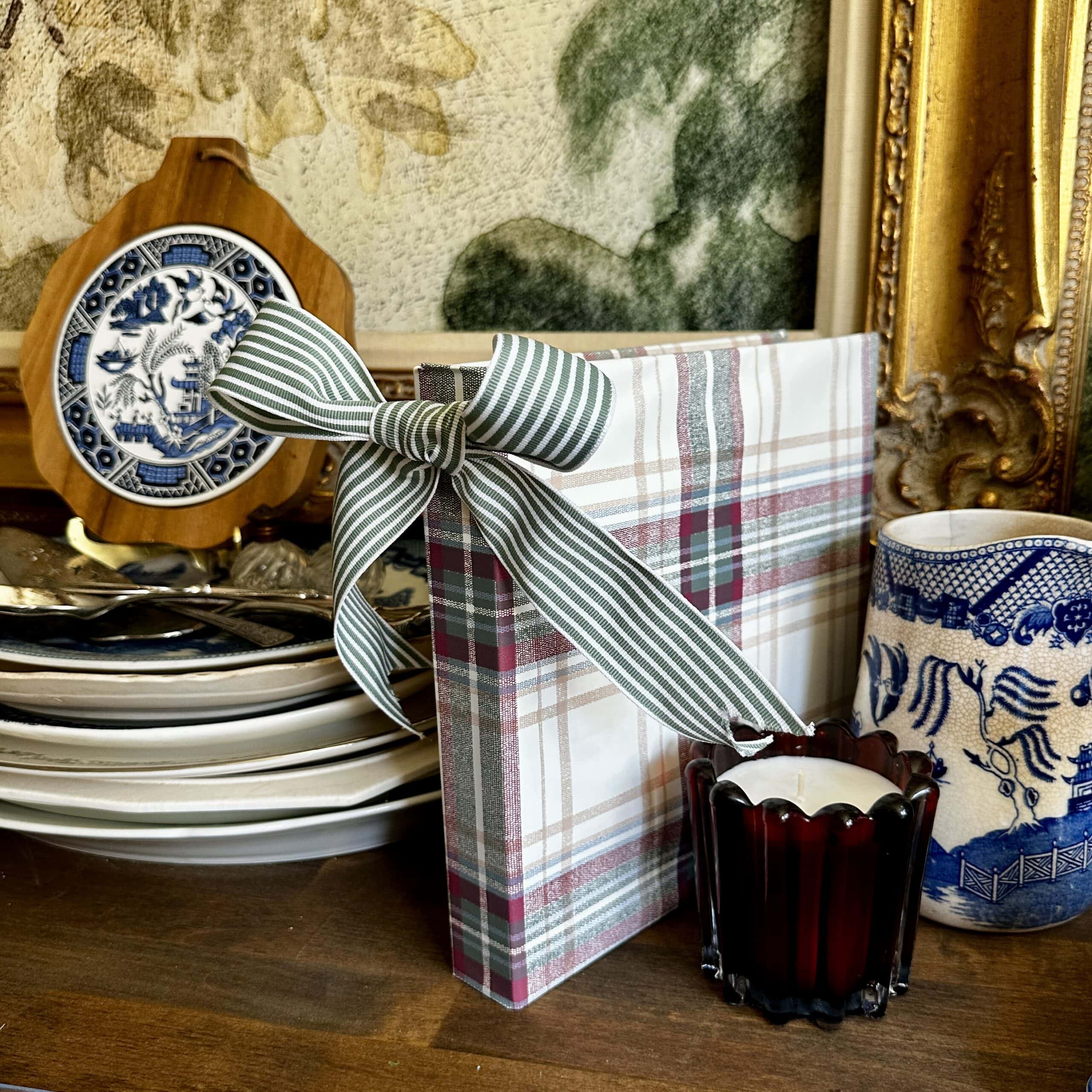 A plaid-wrapped gift with a striped bow sits on a wooden surface near stacked plates, a blue and white dish, a red glass candle holder, and decorative ceramics. A gilded frame and green-patterned wallpaper are in the background.