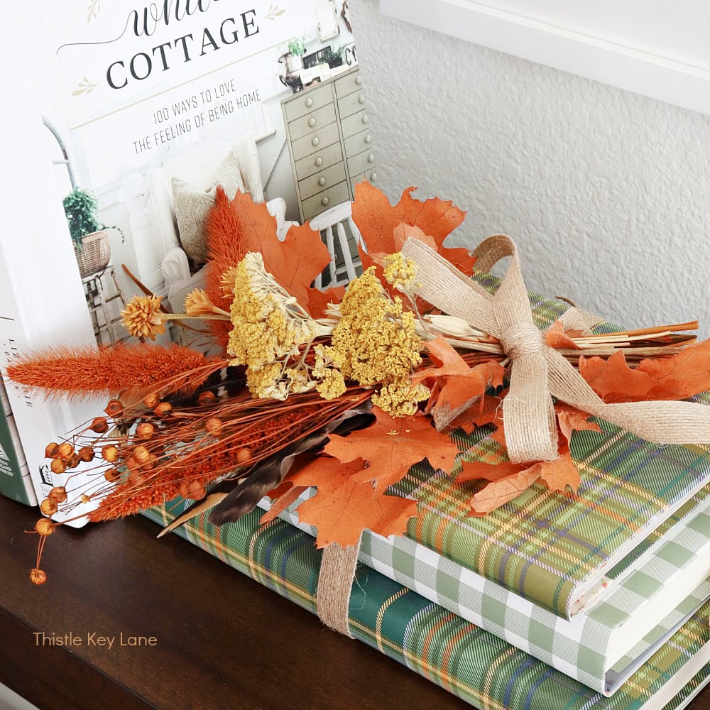 A small fall bouquet with orange leaves, yellow flowers, and twigs tied with burlap ribbon rests on two green plaid books near a home decor magazine.