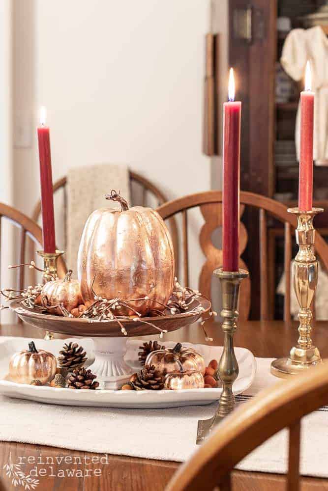 A fall-themed table centerpiece featuring metallic pumpkins, pinecones, berries, and pumpkin crafts on a cake stand, flanked by two lit red candles in brass holders on a wooden dining table.