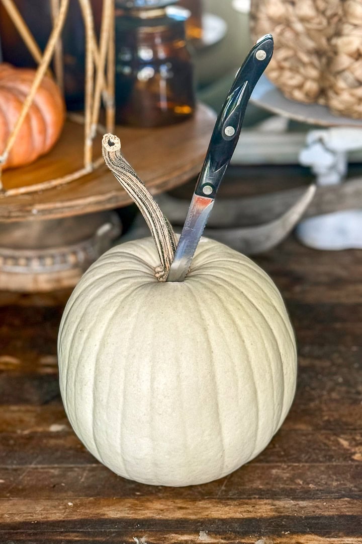 A pale white pumpkin sits on a wooden table with a black-handled knife stuck into its top. Decorative items, including another pumpkin and glass jars, are visible in the blurred background.