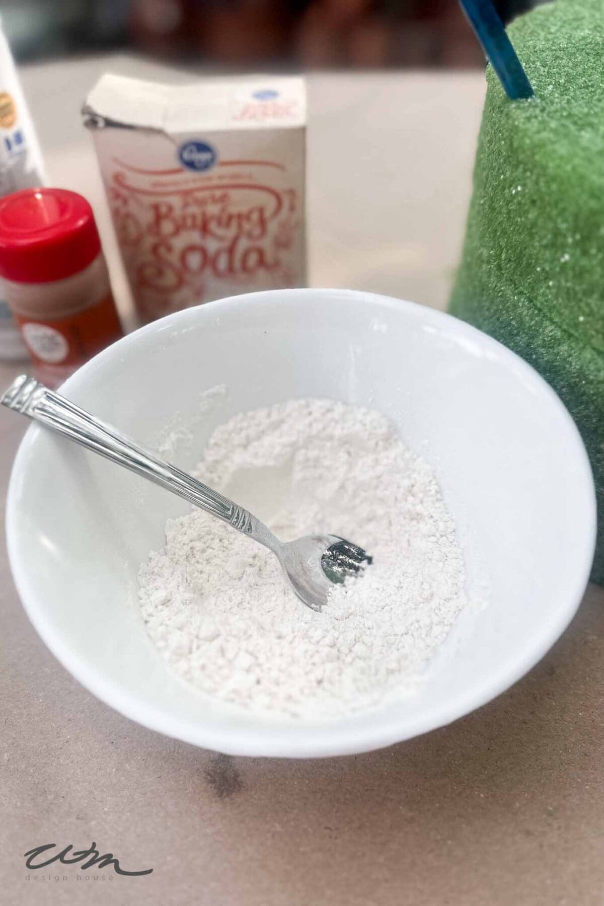 A white bowl with flour and a fork inside sits on a counter. In the background are a box of baking soda, a spice container, and a green foam object.