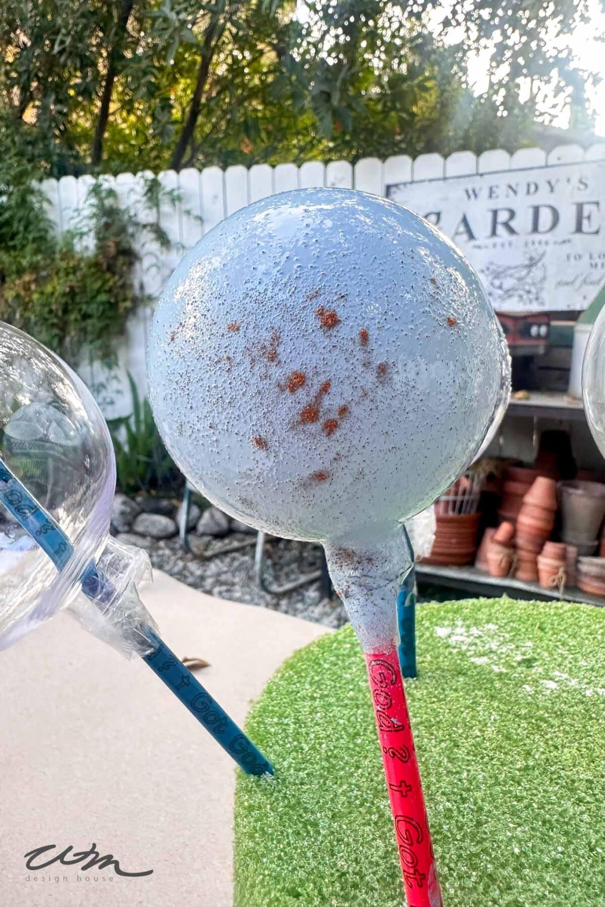 A blue speckled spherical garden ornament on a pink stake stands outdoors on artificial grass, with terra cotta pots, clear globes, and a white fence in the background.