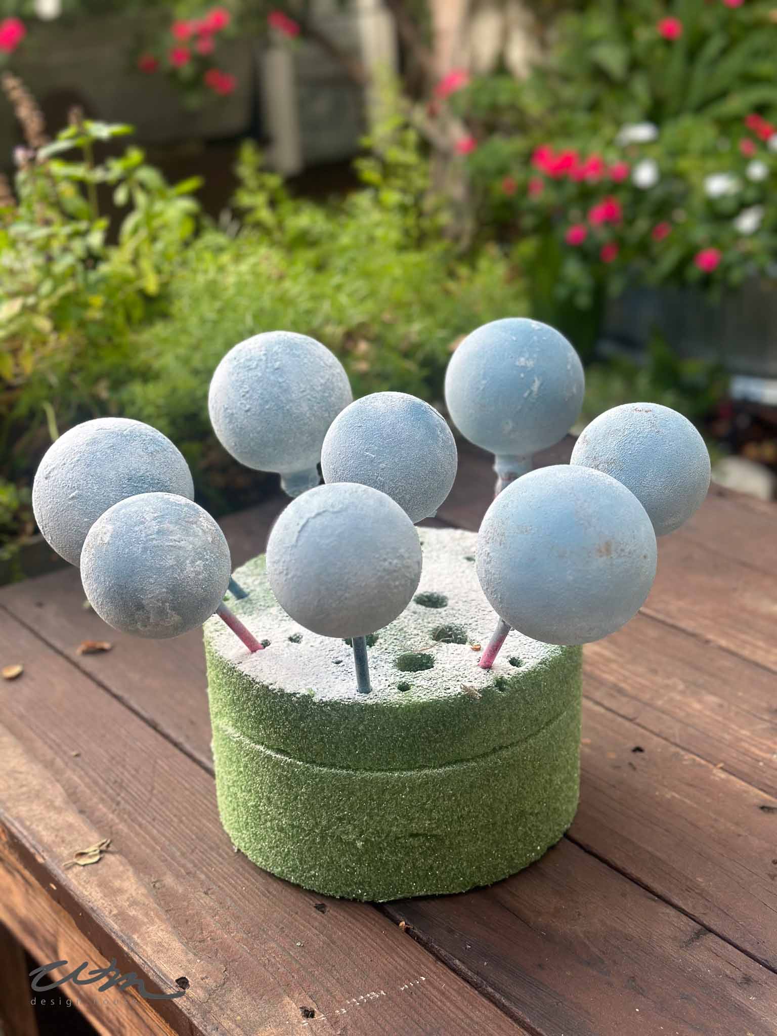 Seven gray spherical objects on sticks are inserted into a round block of green floral foam, placed on a wooden surface outdoors with blurred flowers and greenery in the background.