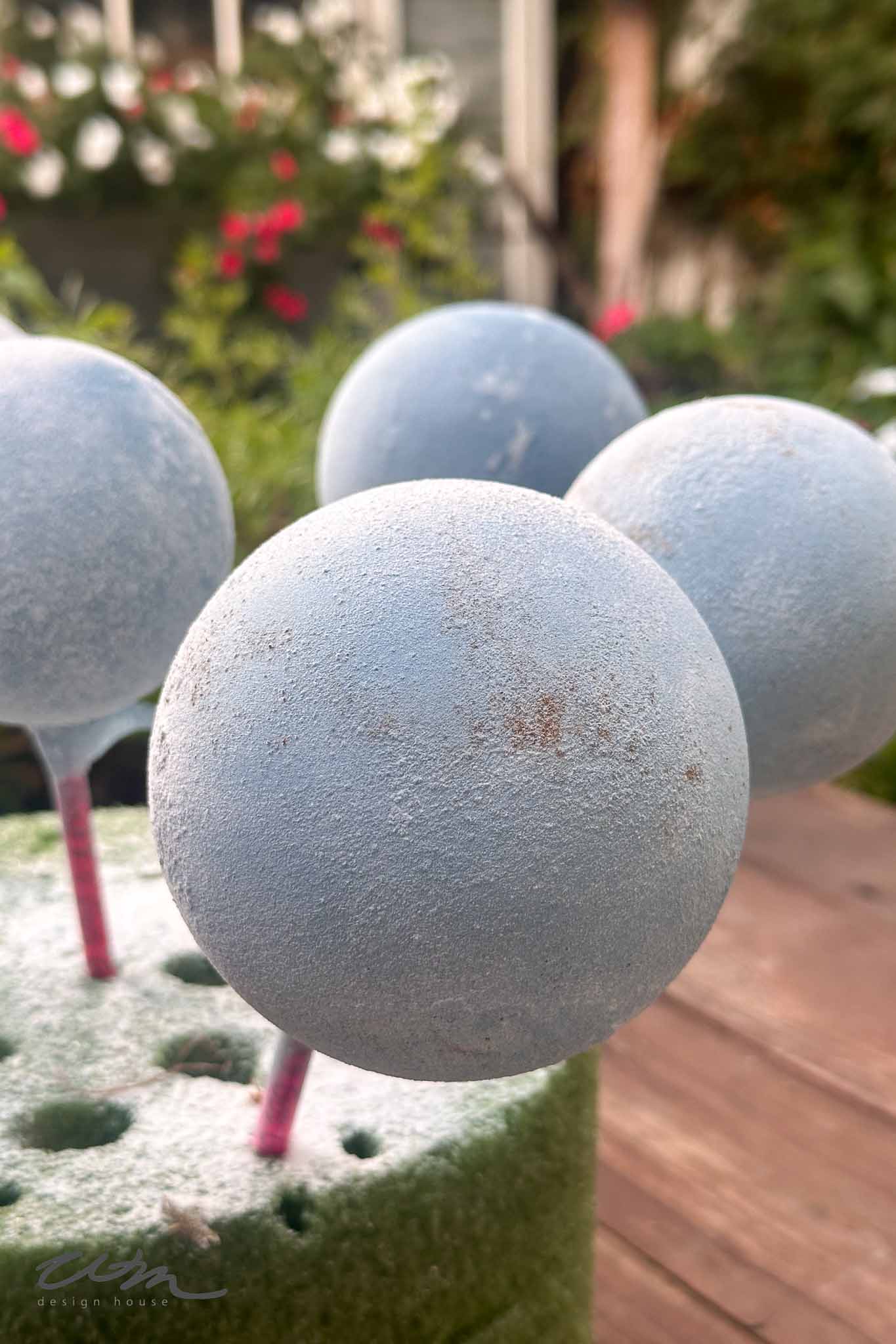 Close-up of three pale blue, round objects on sticks, resembling cake pops or foam balls, standing upright in a green foam block, with a blurred garden background and pink flowers.