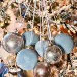 A close-up of several blue and silver Christmas ornaments hanging together in front of a snow-frosted and warmly lit Christmas tree decorated with lights, ribbons, and pine branches.