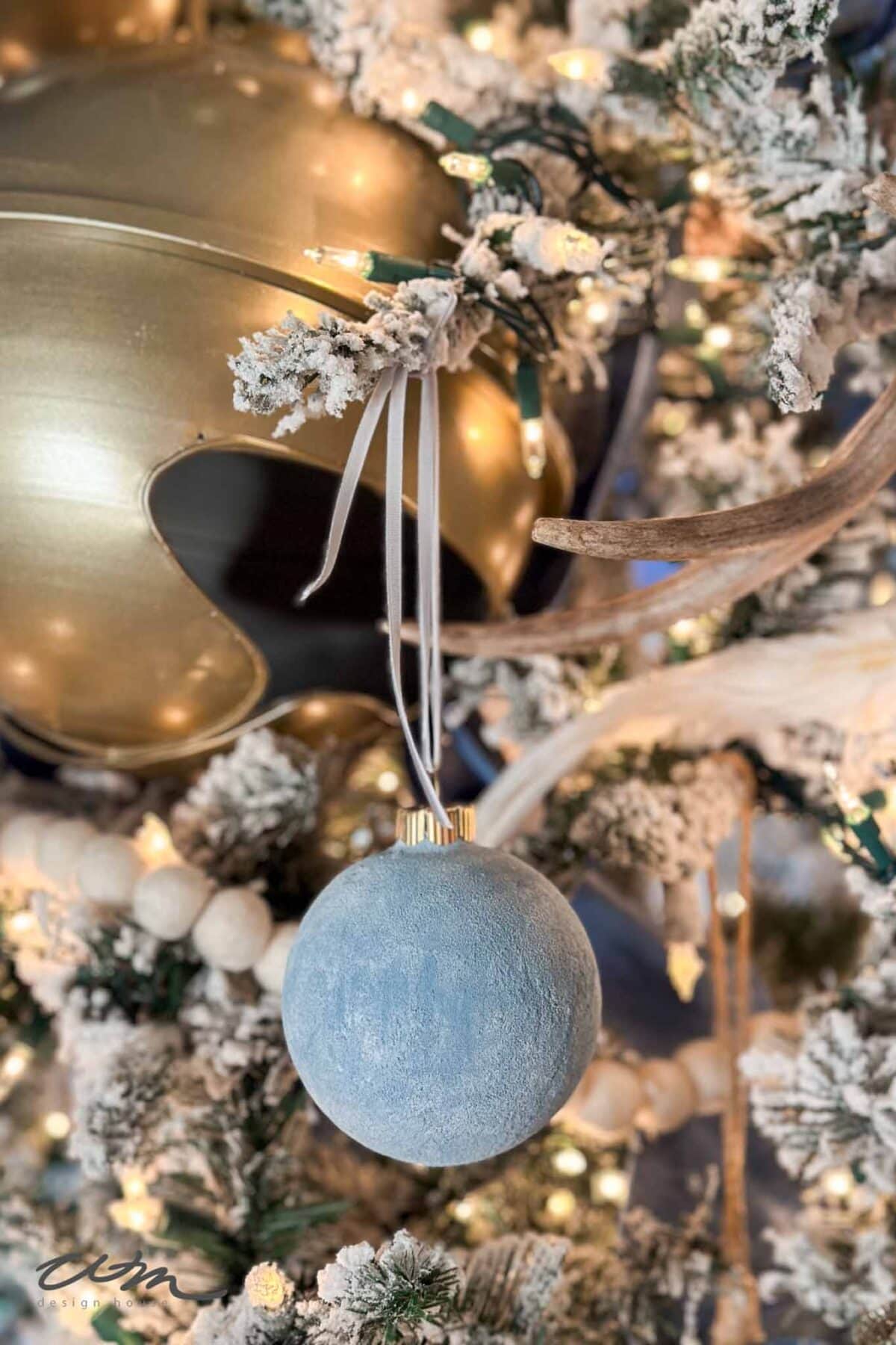 A close-up of a Christmas tree decorated with frosted branches, a blue ornament, white ribbon, gold bauble, and glowing string lights.
