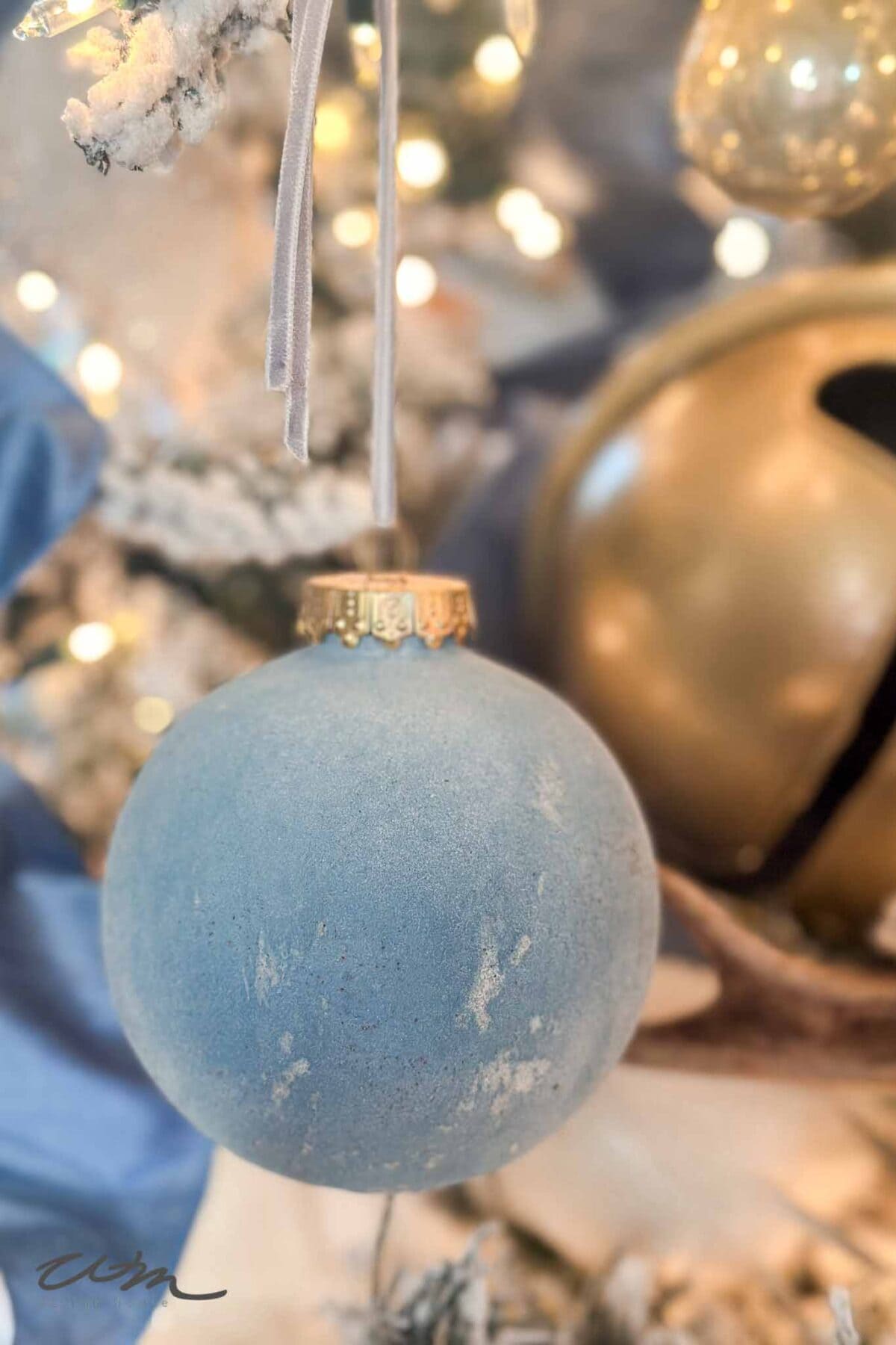 A blue frosted Christmas ornament hangs from a snowy tree branch, with warm white lights and gold decorations softly blurred in the background.