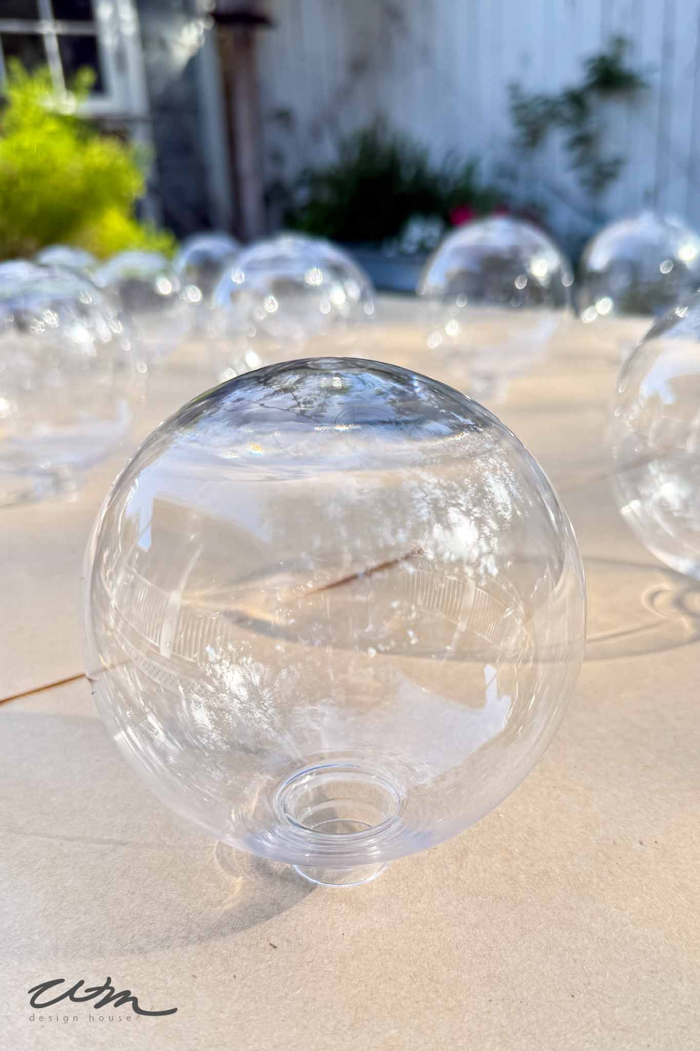 A close-up of a clear, empty plastic ornament ball with an opening at the bottom, sitting on a surface outdoors, surrounded by several similar ornament balls. Sunlight and greenery are visible in the background.