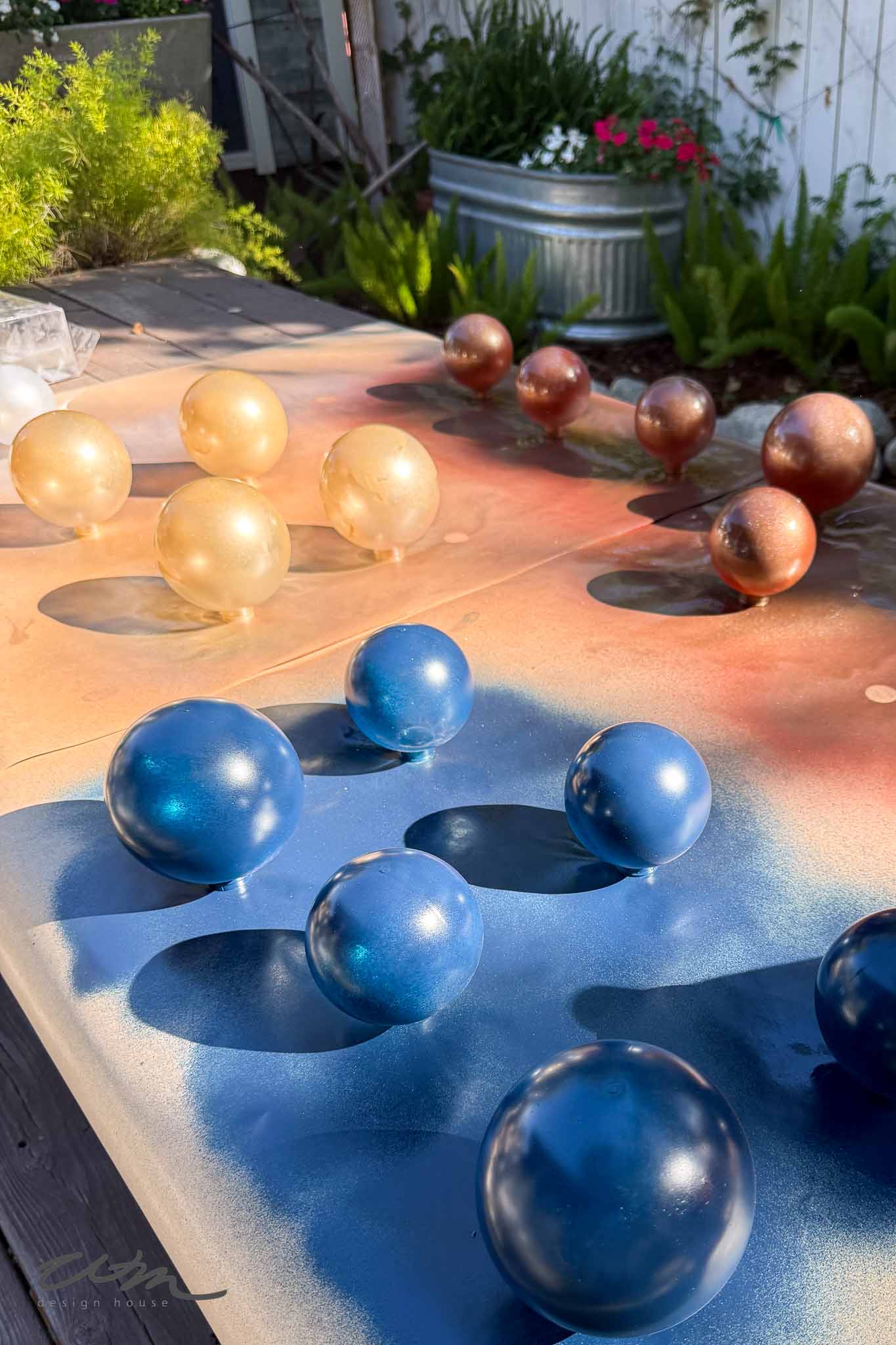 Spheres painted gold, bronze, and blue are drying on a table outdoors, with painted surfaces underneath. There are plants and a metal tub planter in the background. Sunlight casts shadows on the scene.