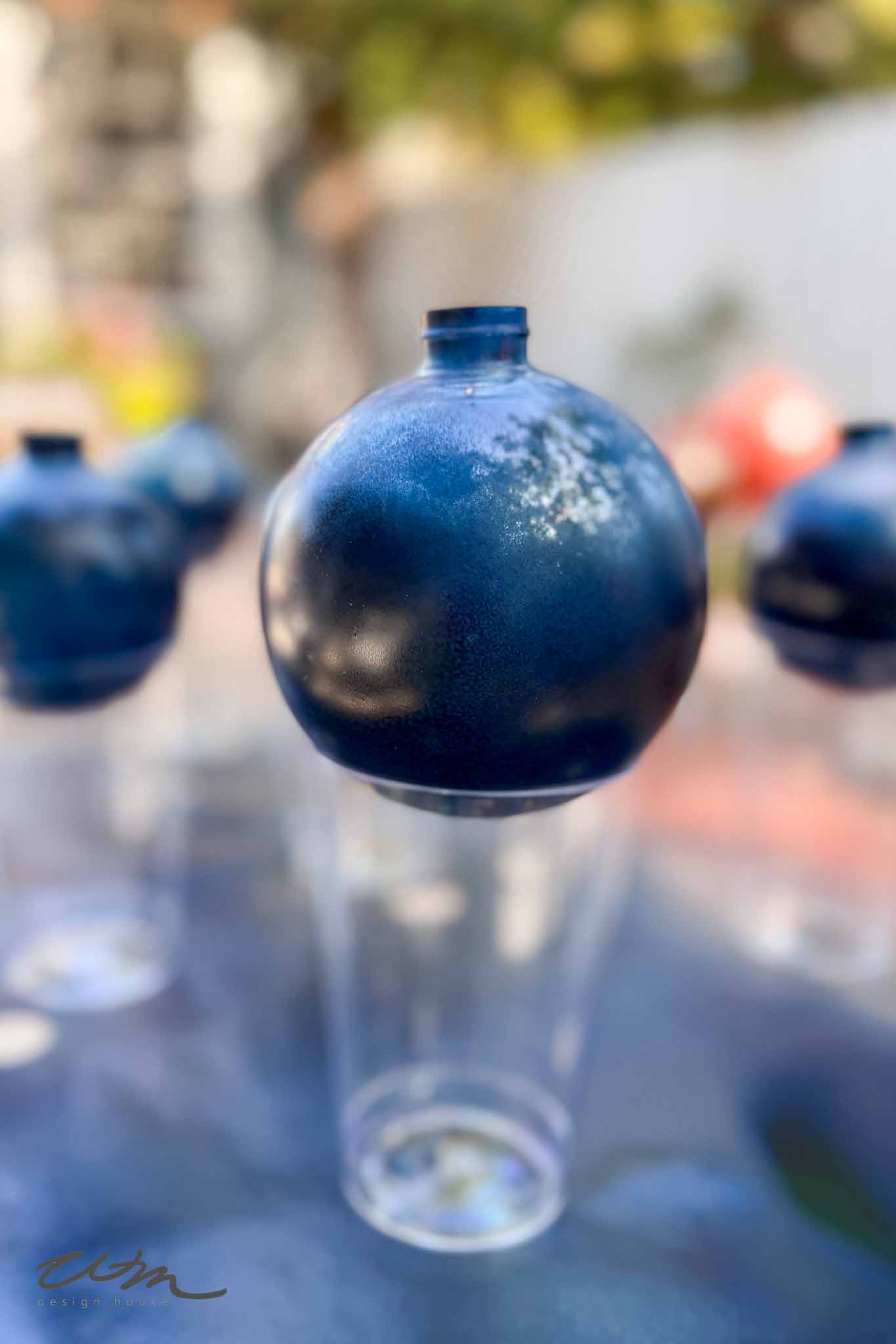 A round object painted dark blue is suspended above a clear plastic cup, with other similar objects and cups blurred in the background. The scene appears to be outdoors.