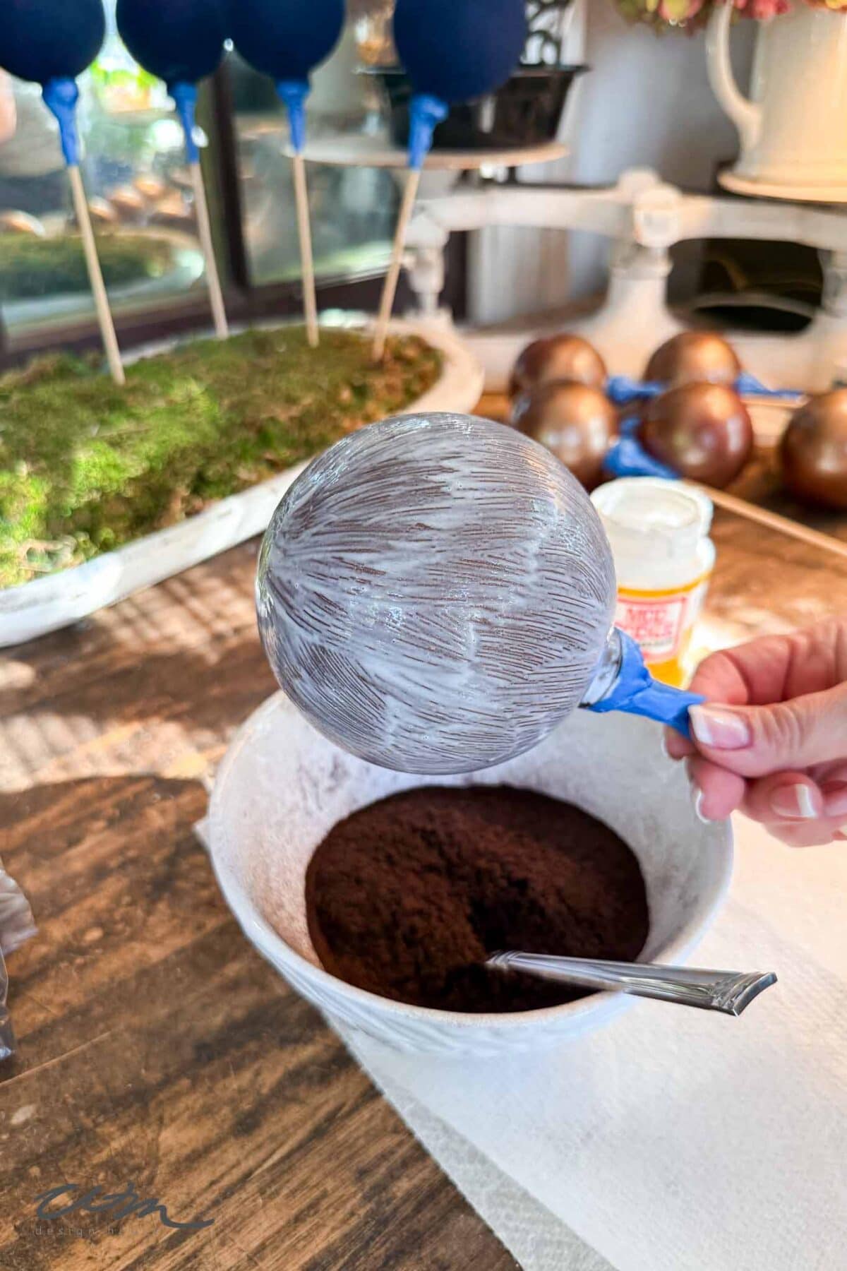 A hand holds a balloon coated with a white substance over a bowl of cocoa powder with a spoon inside; more balloons and chocolate molds are visible in the background on a wooden surface.