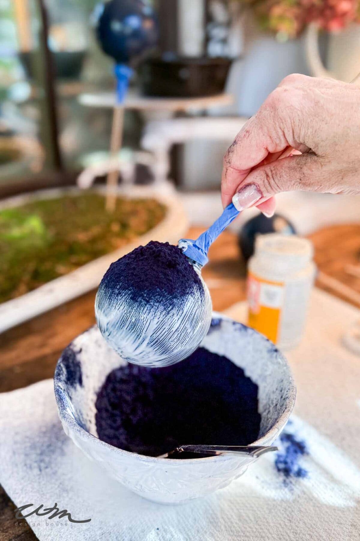 A hand holds a spoon covered in white glue and dipped into a bowl of dark blue powder, likely crafting materials, with some blue powder on the hand and a blurred background.