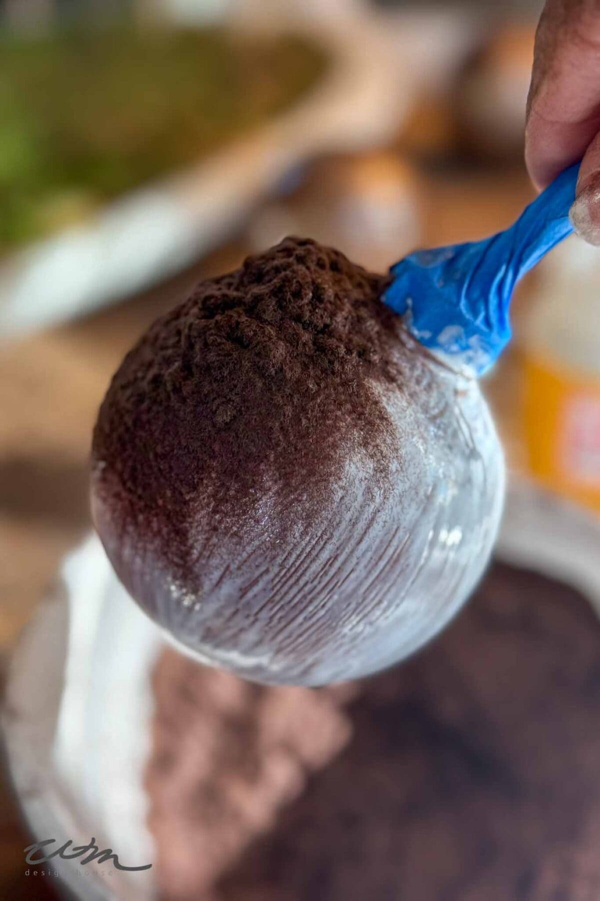 A hand holds a blue plastic spoon scooping up a heaping mound of cocoa powder from a container, with some powder dust visible on the surface.