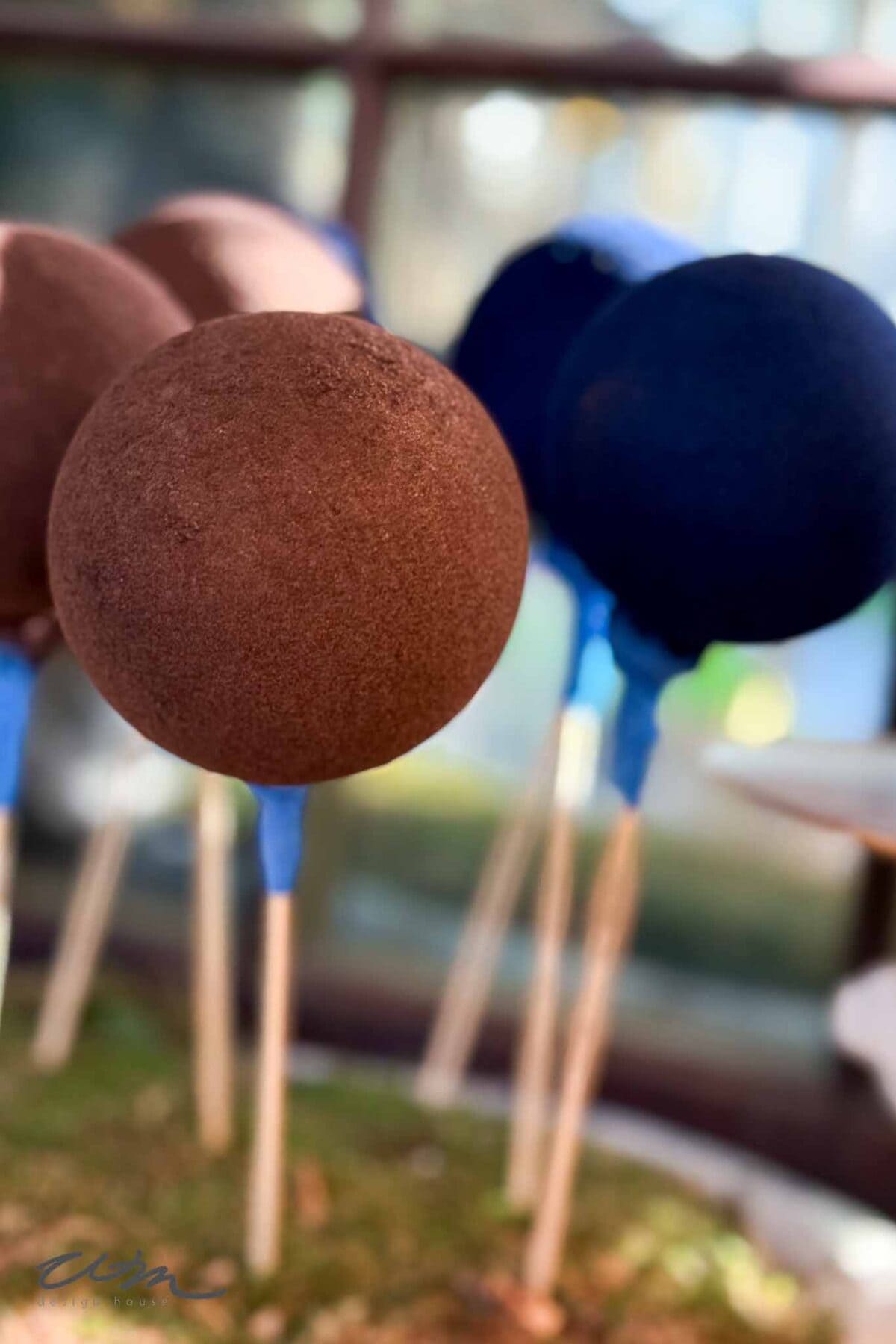 Close-up of round chocolate cake pops on sticks, standing upright. The background is blurred, showing more cake pops and soft natural light through a window.