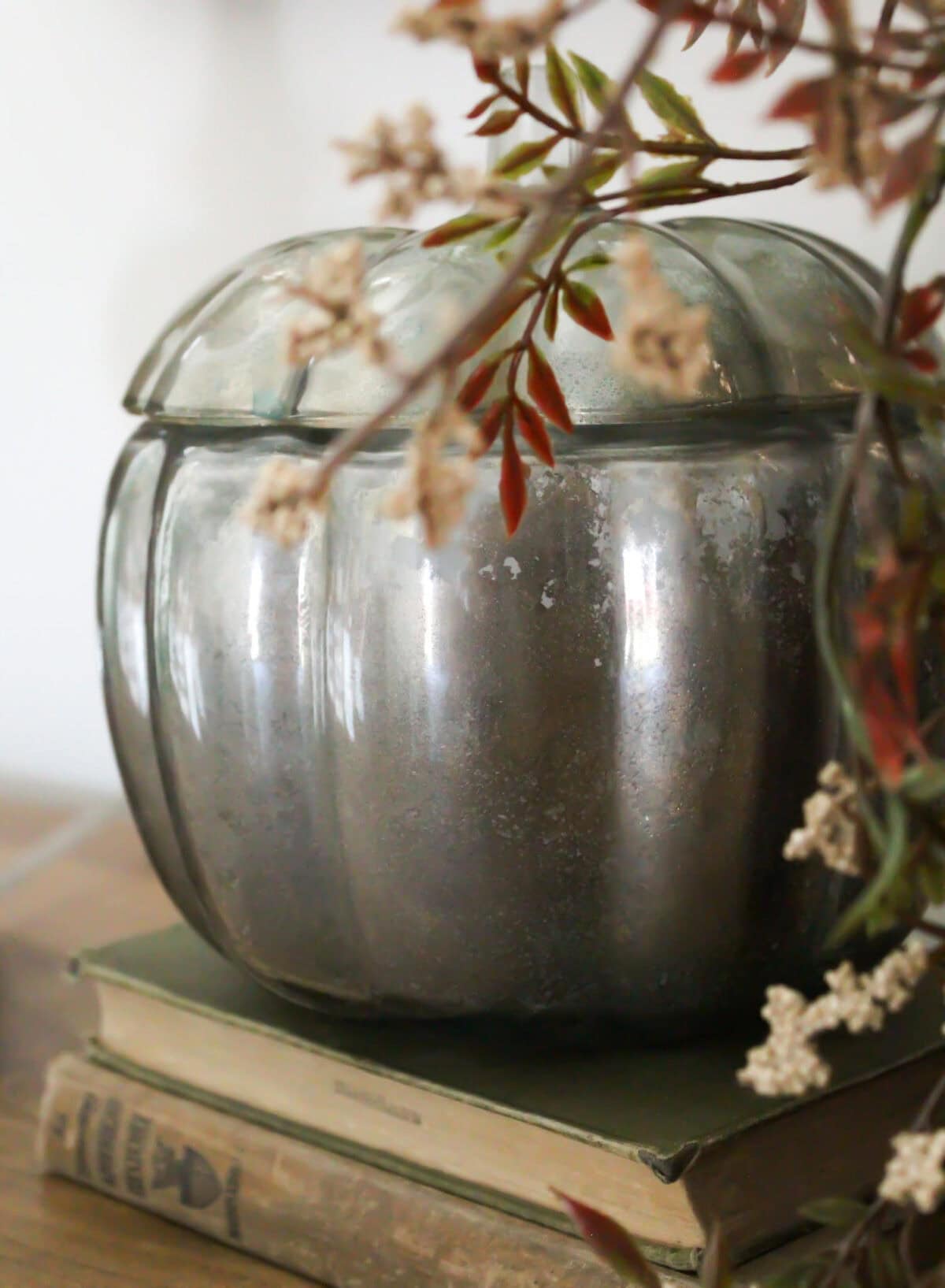 A silver, pumpkin-shaped container—perfect for Pumpkin Crafts—sits on two stacked vintage books, partially framed by delicate branches with small dried flowers.