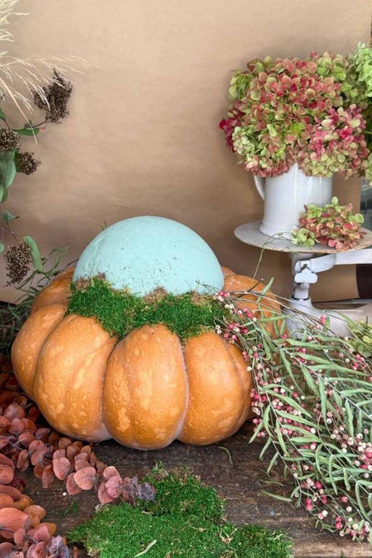 A pumpkin with a light blue dome on top, trimmed with moss, is surrounded by dried and fresh flowers, greenery, and a white vase of hydrangeas against a neutral background.
