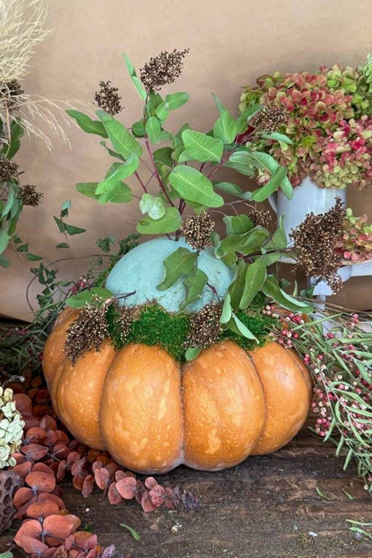 A pumpkin with an arrangement of green leaves, moss, and dried flowers on top, displayed on a rustic wooden surface with more dried plants and flowers around it.