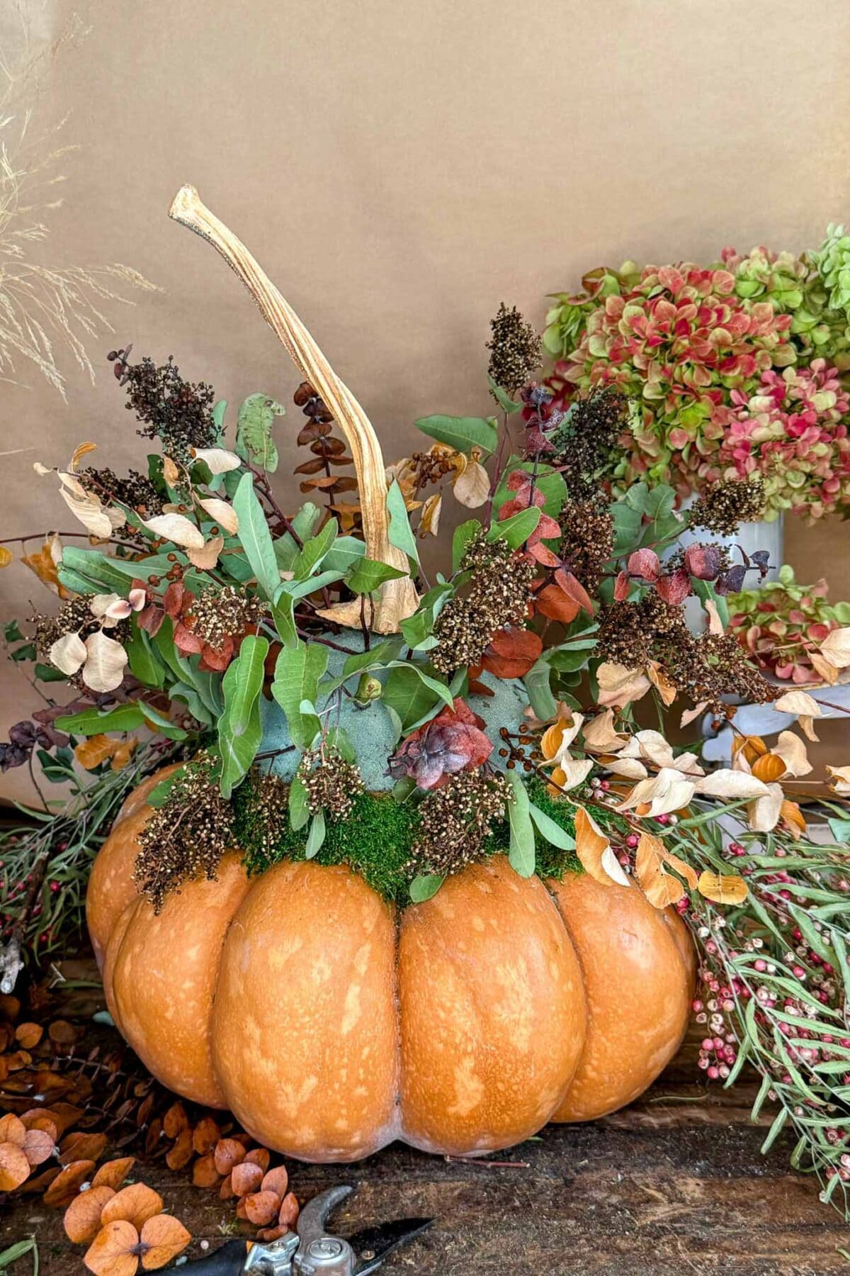 A pumpkin is used as a vase, filled with dried flowers, green leaves, and moss, creating an autumn-themed floral arrangement. Pink and green flowers are in the background on a brown surface.