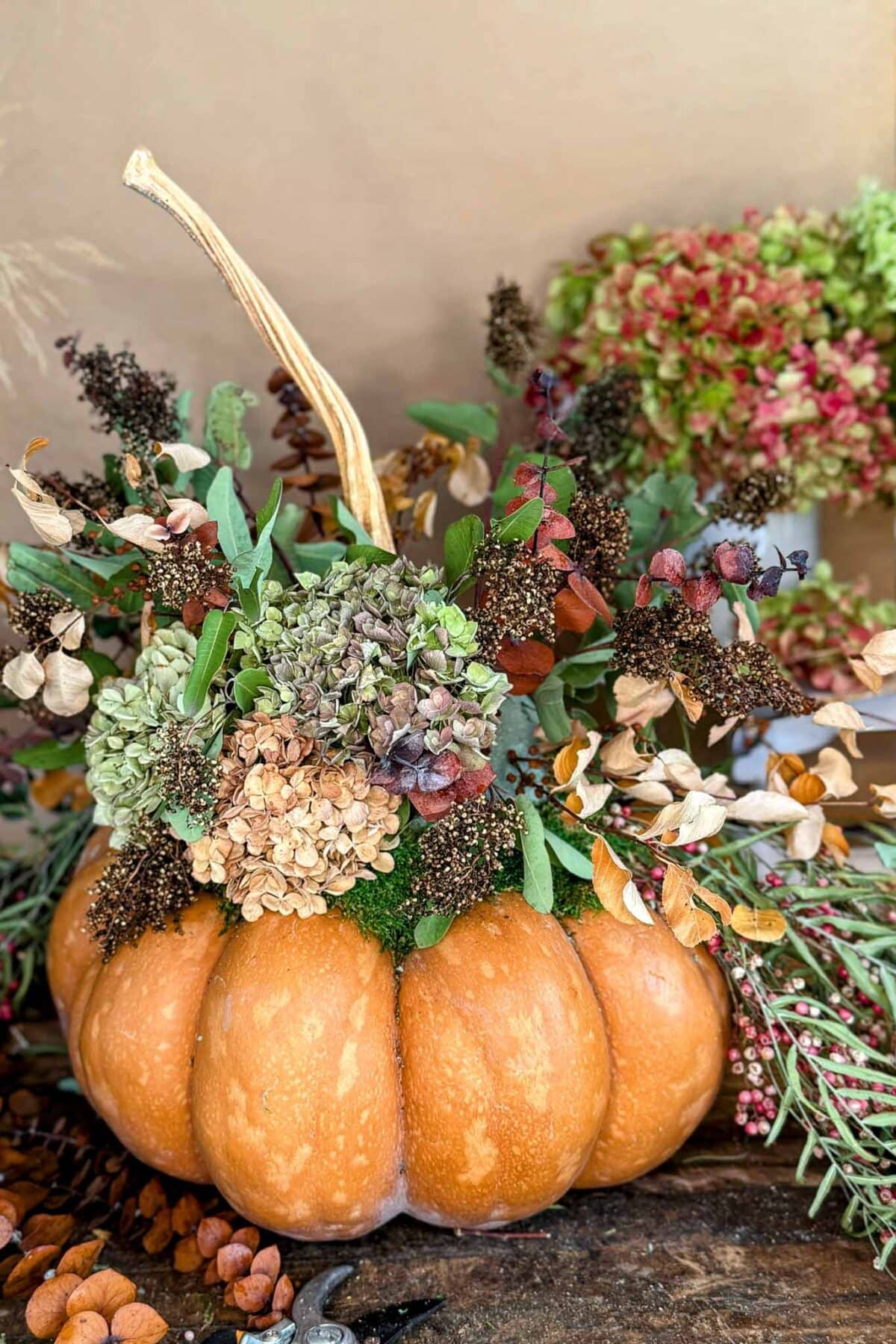 A pumpkin is used as a vase, filled with dried flowers, hydrangeas, leaves, and greenery, creating an autumn-themed floral arrangement. Pinecones and seeds are scattered around the pumpkin.
