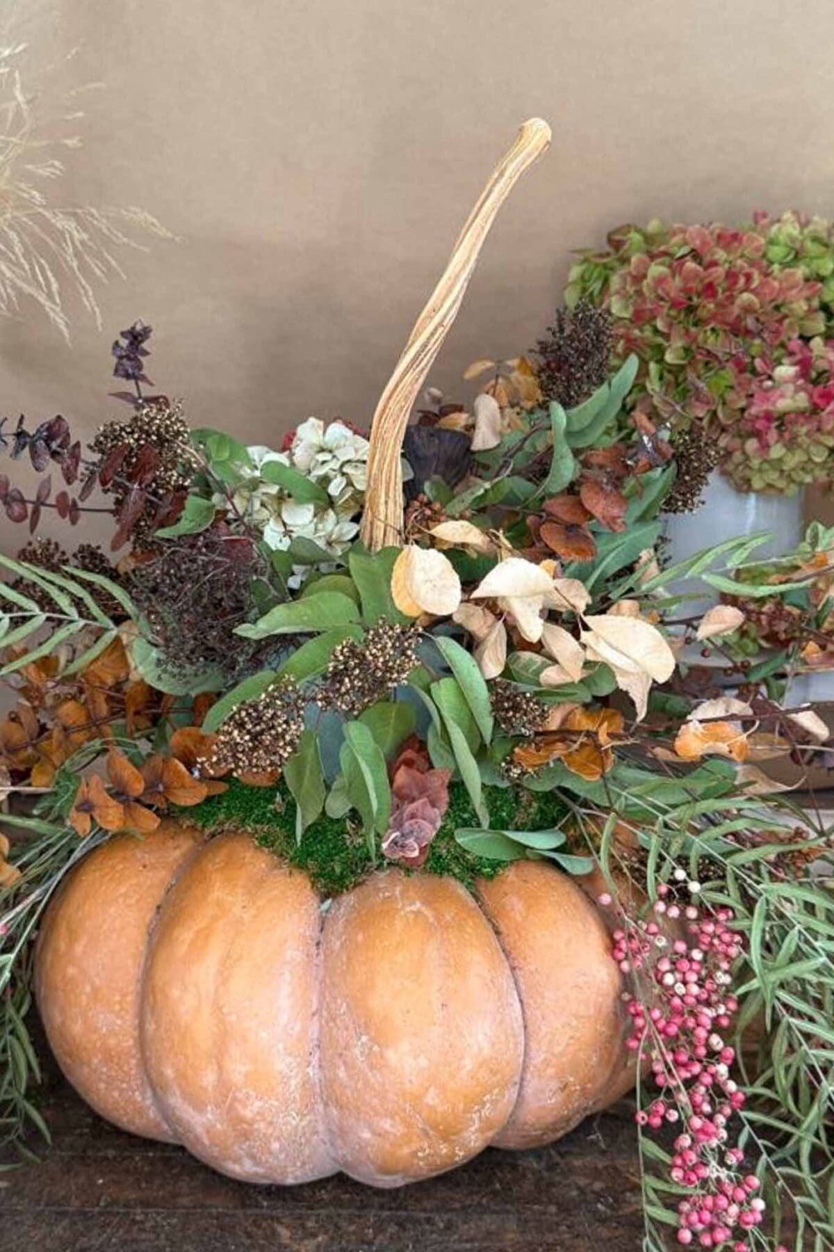 A decorative pumpkin centerpiece filled with assorted dried flowers, leaves, and greenery, creating a rustic autumn arrangement. The pumpkin is placed on a wooden surface with a neutral background.