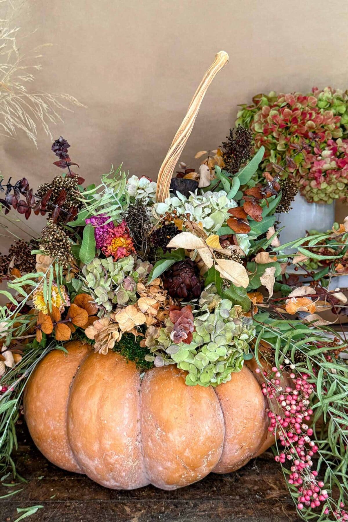 A large orange pumpkin serves as a vase, filled with an arrangement of dried flowers, leaves, and greenery. The display has a rustic, autumnal feel, with the pumpkin stem curving gracefully upward.