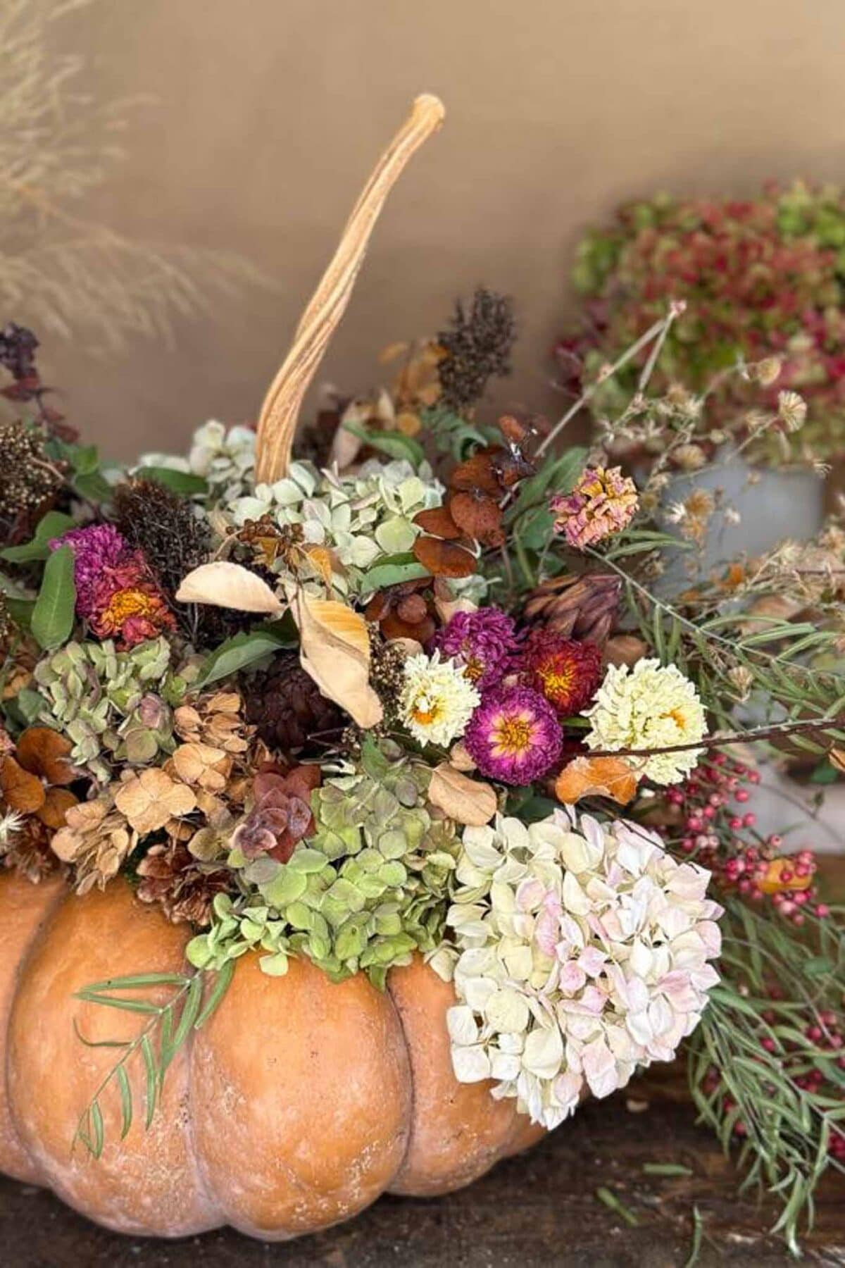 A large pumpkin serves as a vase, filled with assorted dried and fresh flowers in shades of purple, pink, cream, and green, creating an autumn-themed floral arrangement.