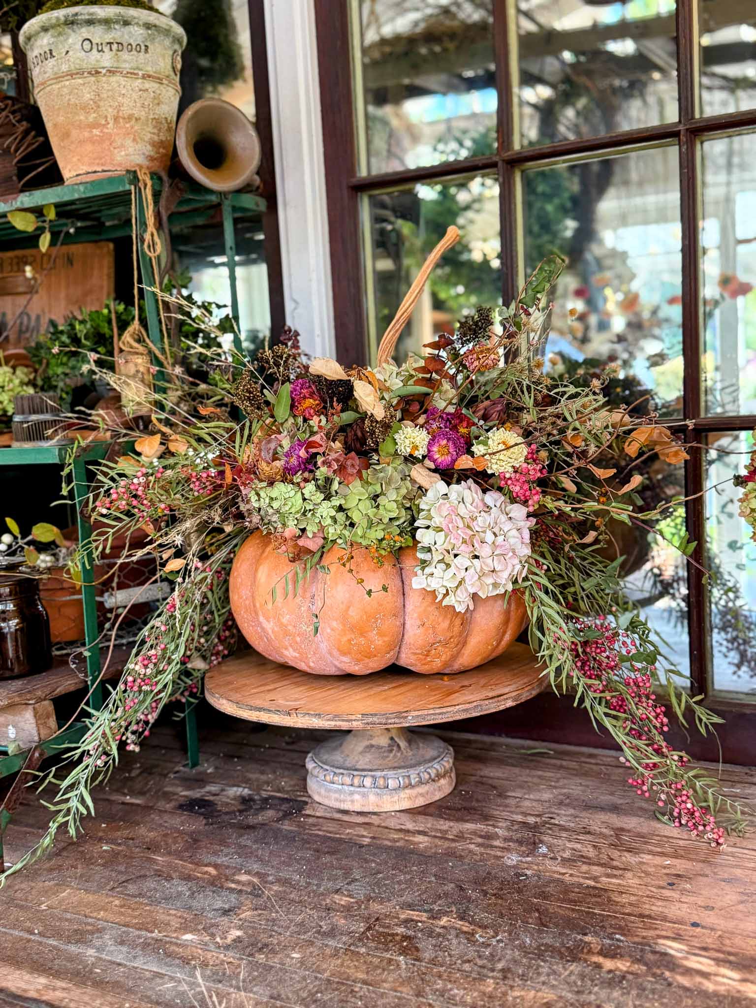A large pumpkin is used as a vase to create a floral arrangement with colorful dried wildflowers, hydrangeas, and greenery, and displayed on a wooden pedestal. The arrangement sits on a rustic wooden table in front of a window and shelves with garden items.