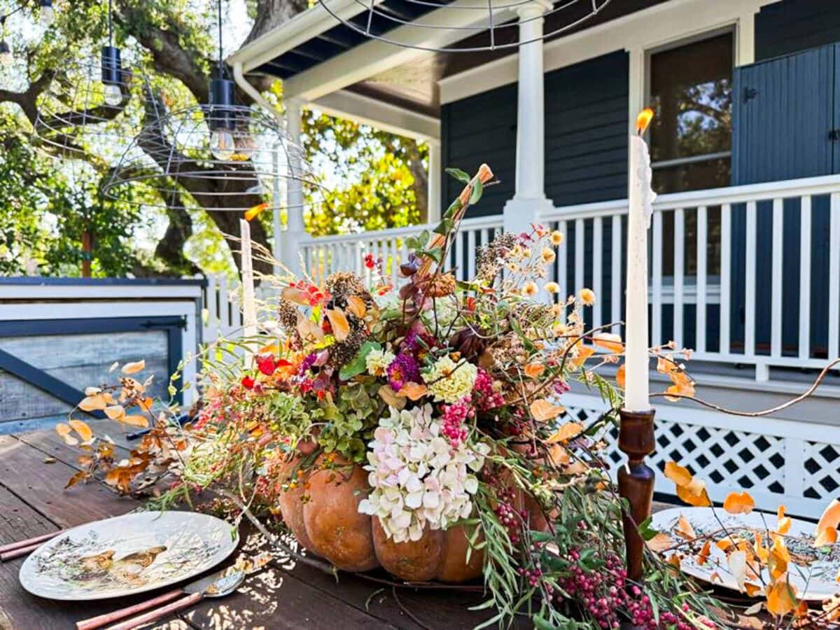A rustic outdoor table displays a large pumpkin centerpiece filled with colorful autumn flowers and leaves, with plates, cutlery, and a lit candle beside it. A house with dark shutters and a porch is in the background.
