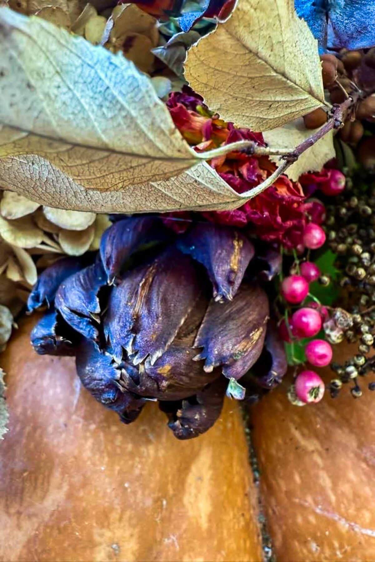 Close-up of a dried artichoke surrounded by brown and purple leaves, red berries, and dried flowers, creating an autumnal, textured arrangement on a wooden surface.
