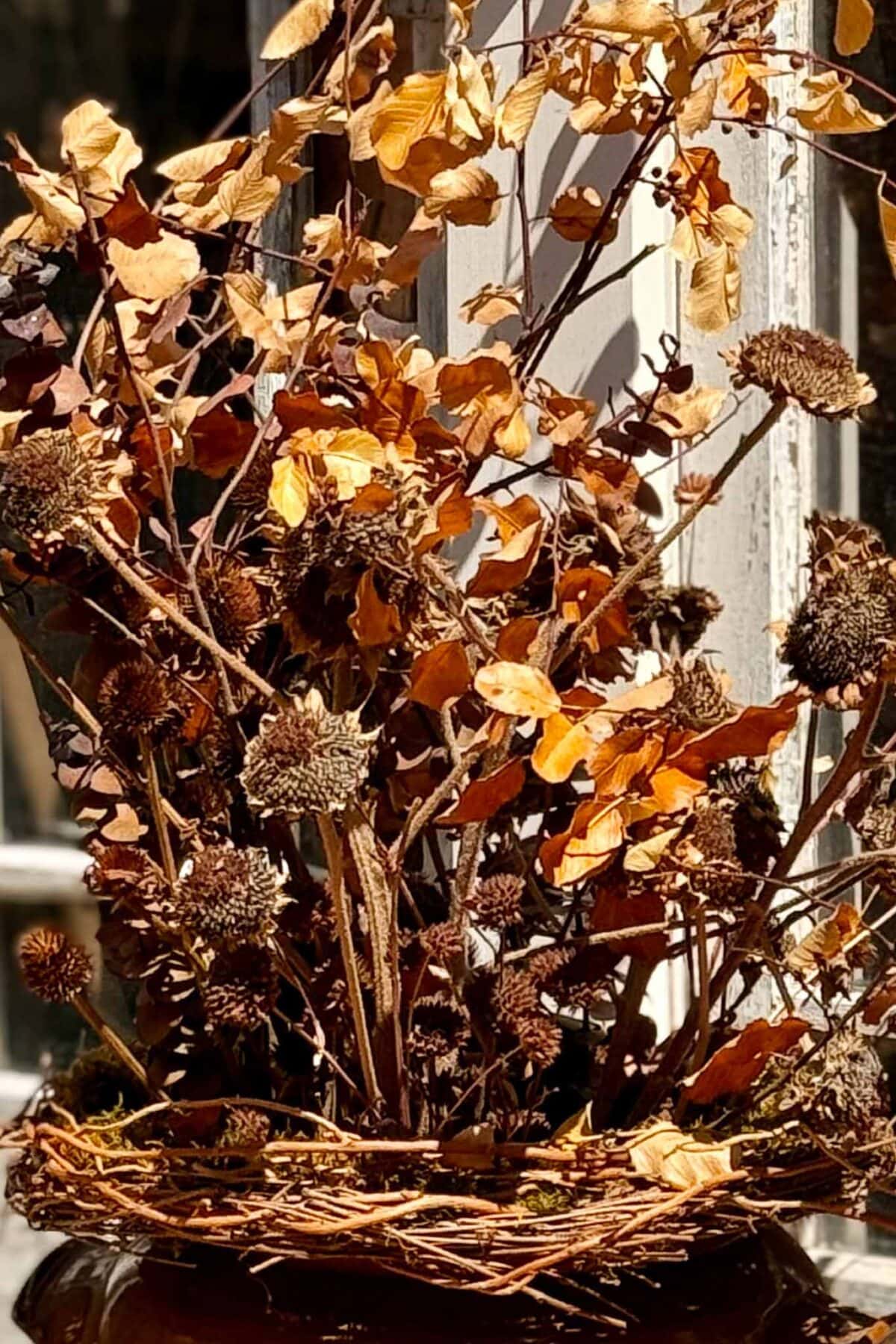 A close-up of a basket filled with dried brown and yellow leaves and flowers sits in sunlight near a rustic window, creating a charming dried flower pumpkin centerpiece.