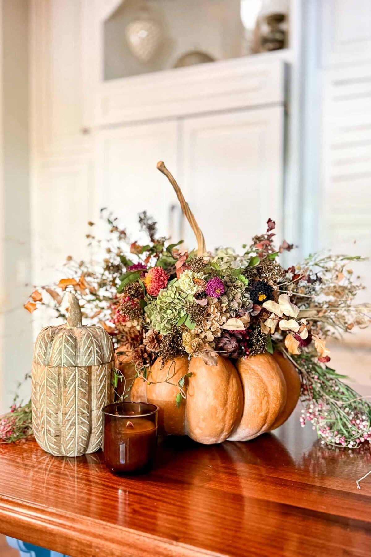 A Dried Flower Pumpkin Centerpiece sits on a wooden table next to a carved decorative pumpkin and a small brown candle, with a cozy kitchen background.