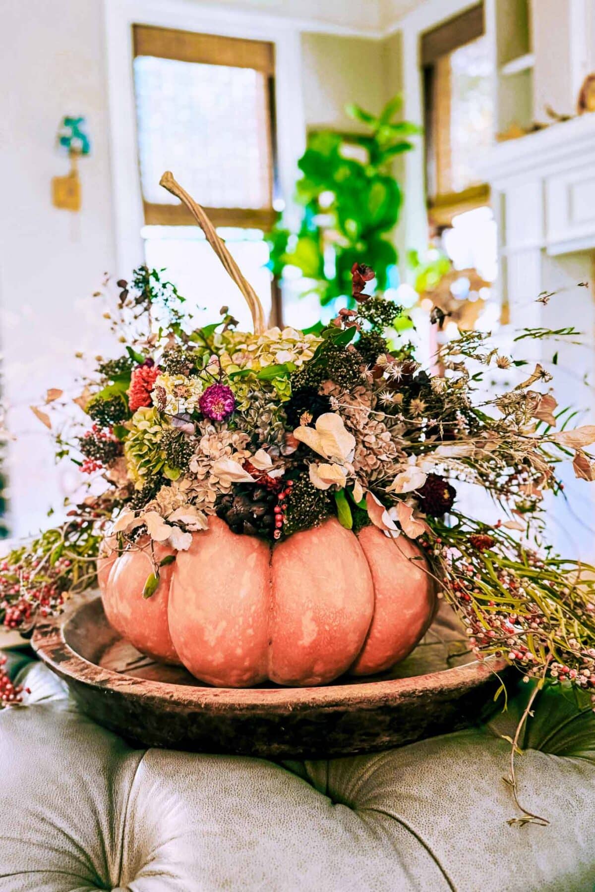 A Dried Flower Pumpkin Centerpiece features a pink pumpkin filled with dried flowers, leaves, and berries, beautifully arranged in a rustic wooden bowl on a table in a bright, cozy room.