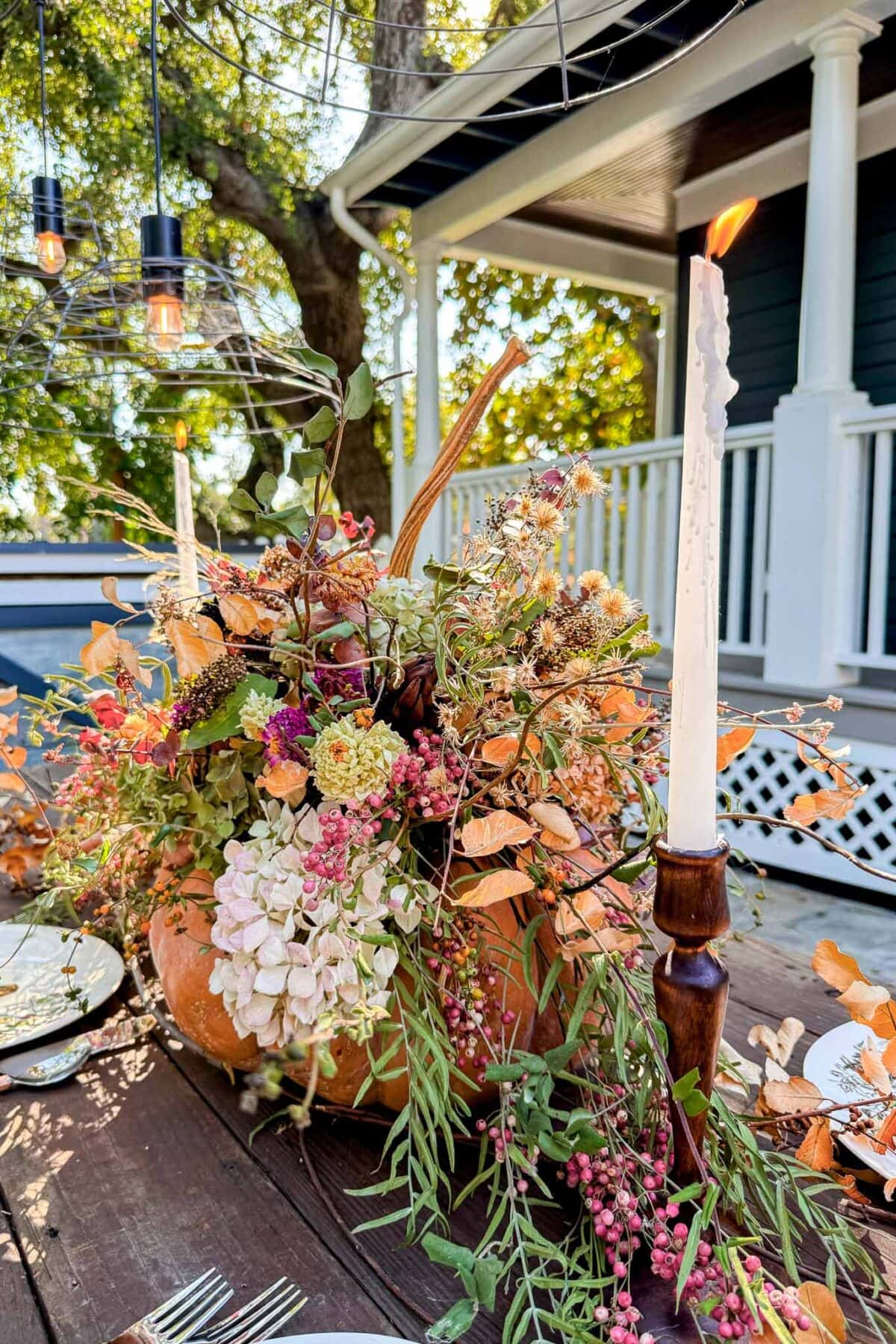 A rustic outdoor table features a dried flower pumpkin centerpiece filled with autumn blooms and greenery, flanked by lit taper candles. Warm sunlight filters through trees and a porch is visible in the background.