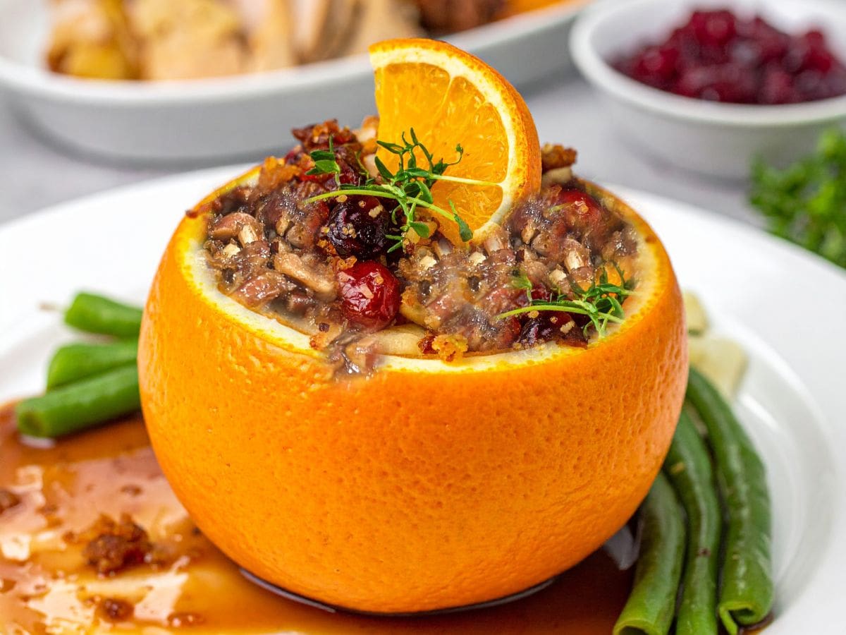 A hollowed orange filled with stuffing, topped with cranberries, herbs, and an orange slice, sits on a white plate with green beans and brown sauce—these Thanksgiving Sweet Potato Orange Cups make a festive addition, with cranberry sauce in the background.