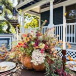 A rustic outdoor table features a Dried Flower Pumpkin Centerpiece, overflowing with vibrant blooms and foliage. Plates and utensils are set nearby, with a candle and a white house with blue shutters in the background.